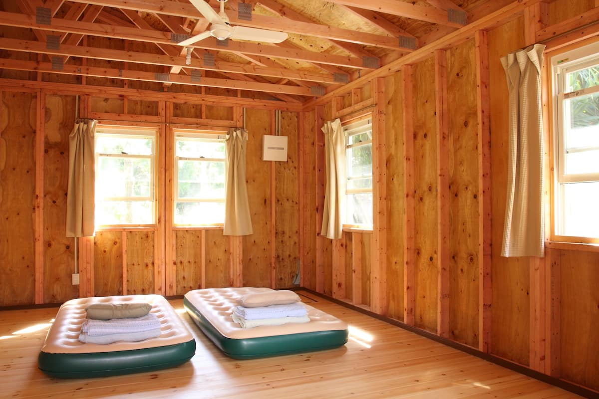 Une pièce aux murs et au plancher en bois, avec deux lits simples dotés de matelas et des rideaux aux fenêtres.