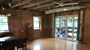 Una habitación amplia con pisos de madera, una mesa de comedor con sillas, un pequeño estante y un ventanal corredizo que da a un espacio exterior con plantas en macetas.