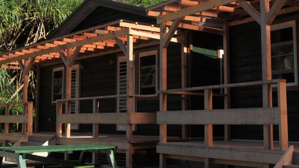 Una cabaña de madera con un porche y mesas de picnic, rodeada de exuberante vegetación tropical y una montaña al fondo.