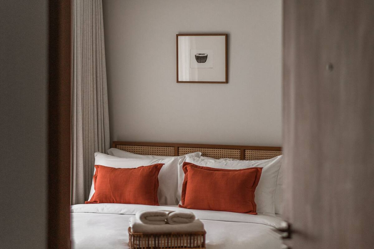 A neatly made bed with white and orange pillows, a wooden headboard, and a framed picture on the wall.