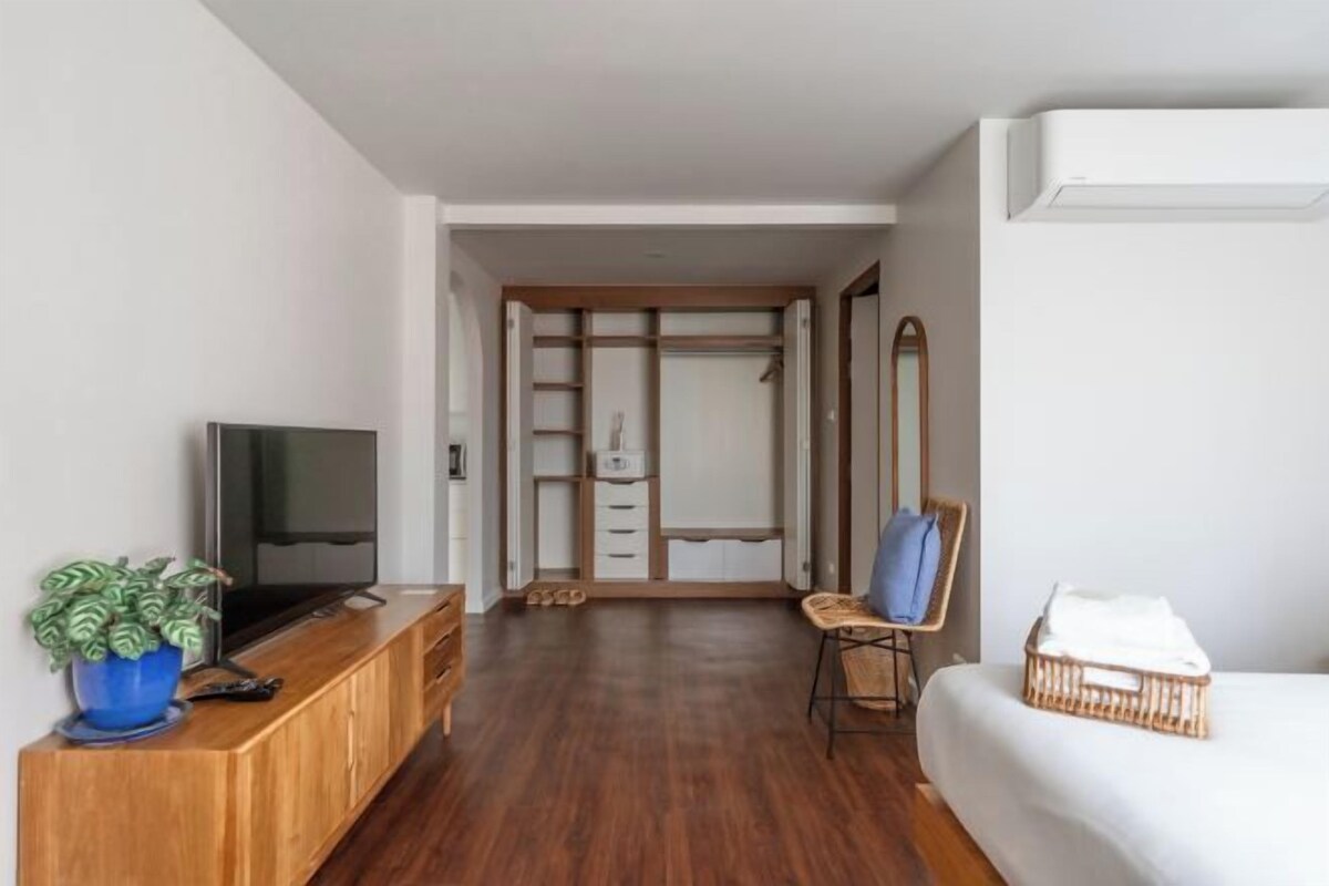 A modern room with a wooden floor, a television on a wooden cabinet, a plant, a bed with a white comforter, and a wardrobe with sliding doors.