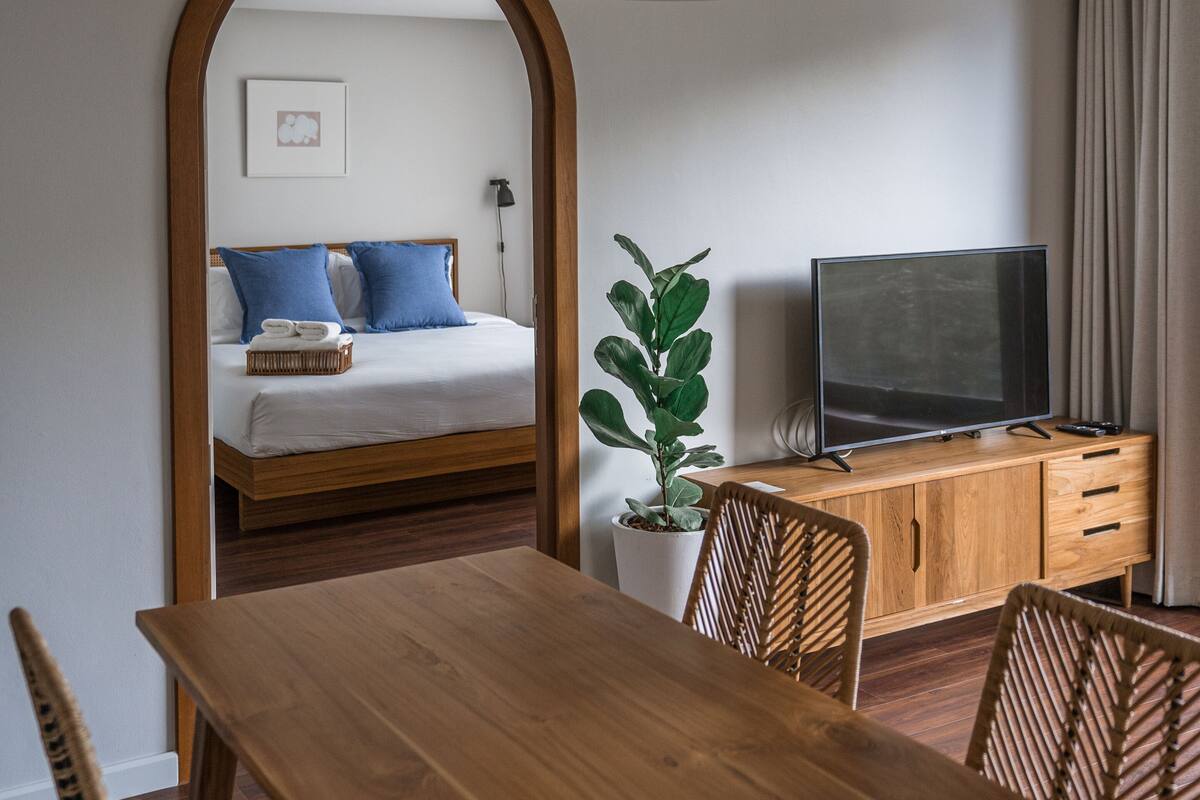 A modern hotel room with a wooden dining table, chairs, a TV, and a bed with blue pillows.