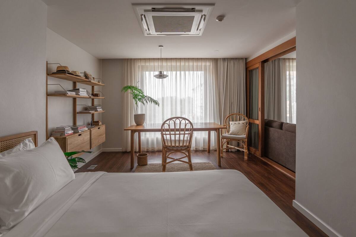 A hotel room with a bed, a desk with a chair, a bookshelf, a potted plant, and a large window with curtains.