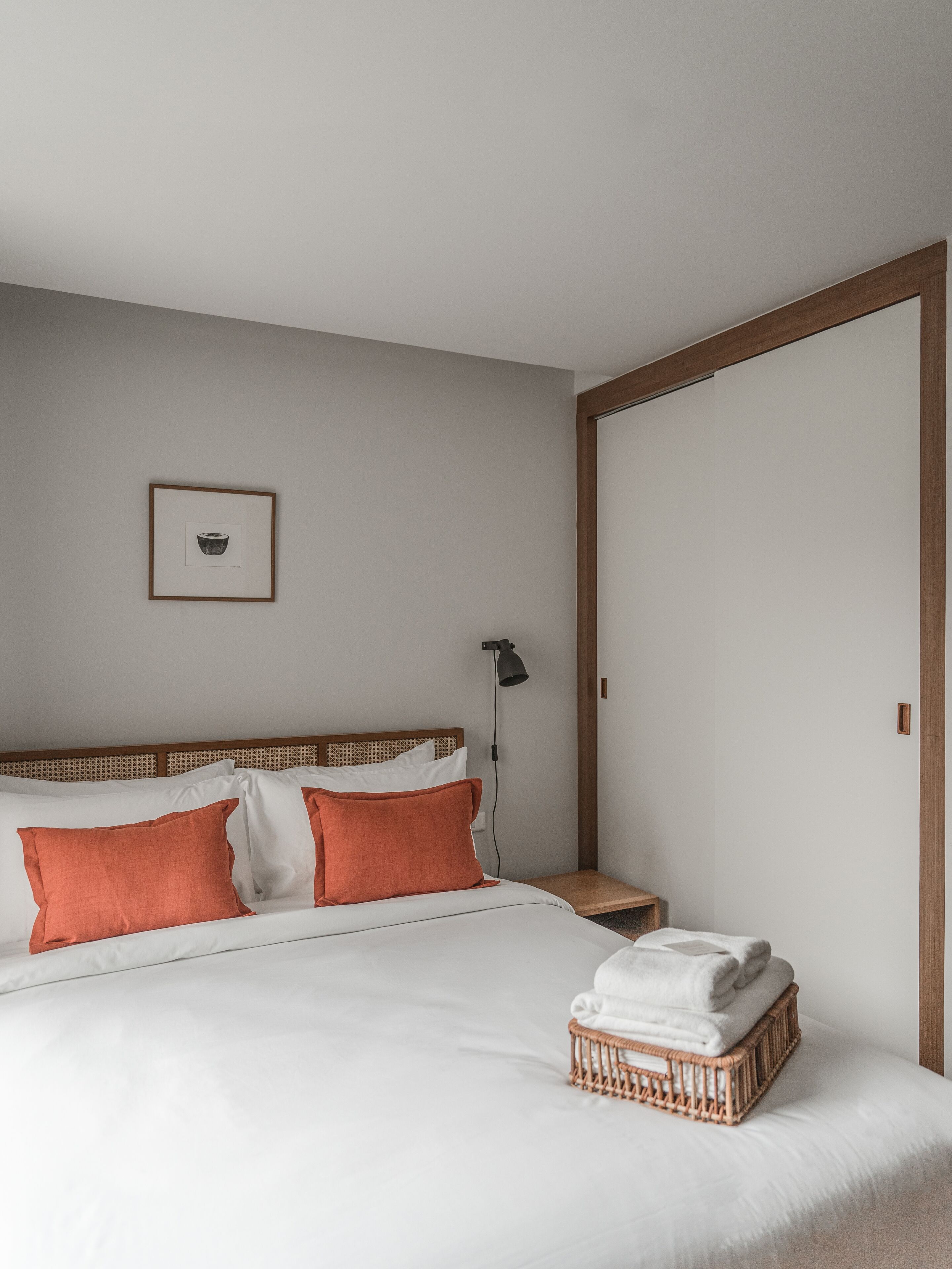 Una cama bien hecha con almohadas blancas y anaranjadas, una mesita de noche con una lámpara y una cesta de mimbre sobre la cama.