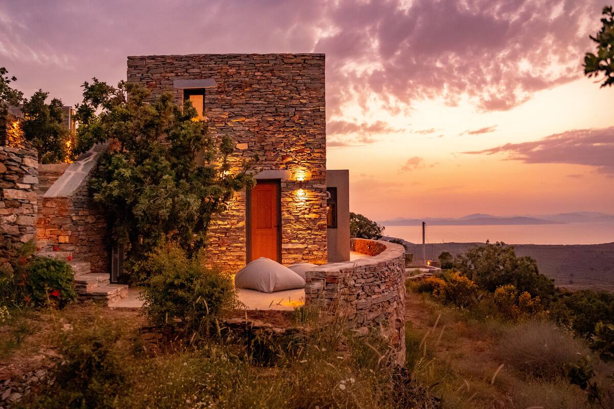 A stone building with a wooden door and a small window, surrounded by vegetation and a sunset view.
