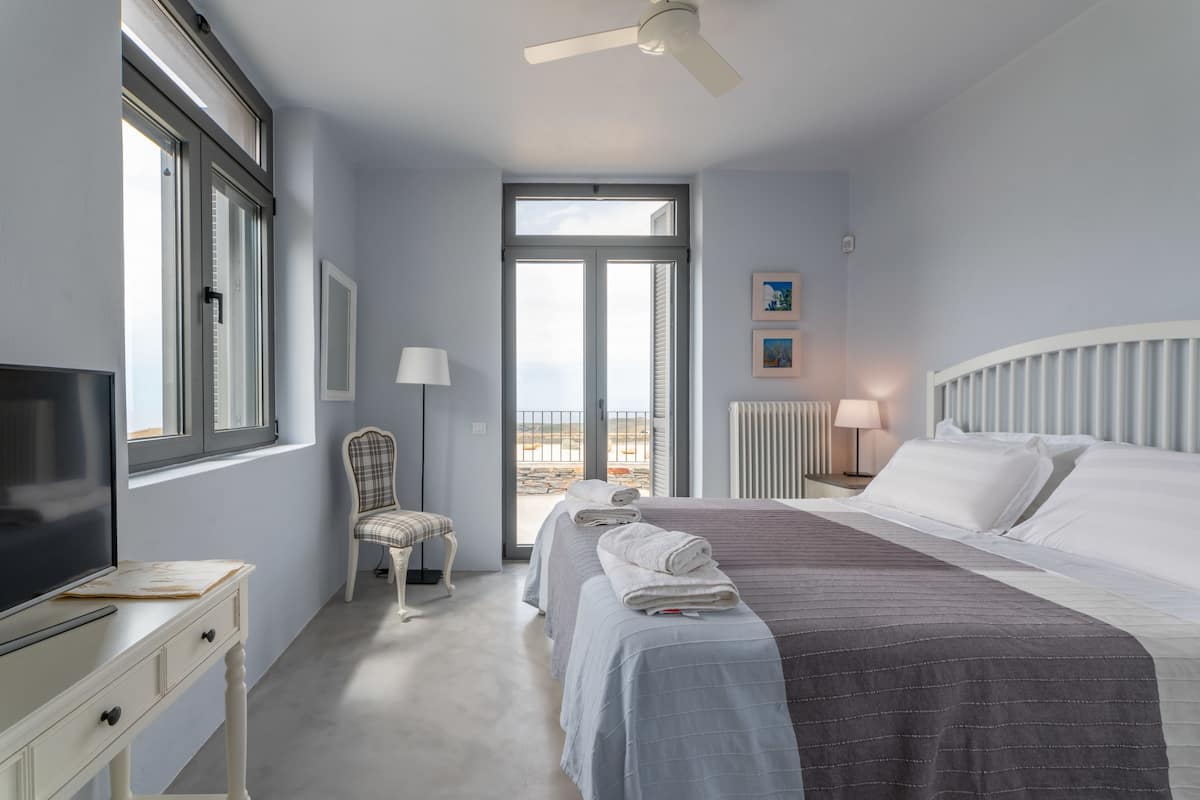 A bedroom with a bed, a television, a chair, a window, and a view of the outdoors.