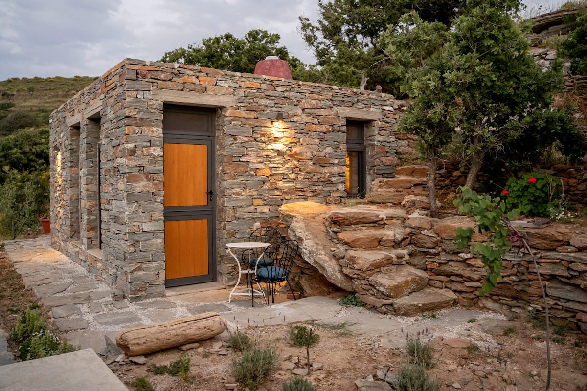 A stone building with a wooden door and window, a small table with chairs, and a stone pathway.