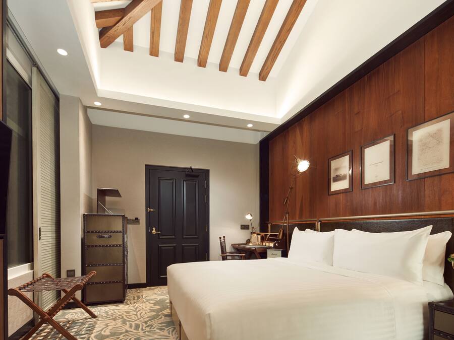A hotel room with a large bed, wooden ceiling beams, a desk with a chair, a wardrobe, and framed pictures on the wall.