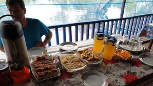 A breakfast table with various food items and a kettle, set on a balcony with a view of water. - Jungle Reps (Iquitos)