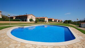 A swimming pool with a stone border, surrounded by a well-maintained lawn and a row of buildings in the background.