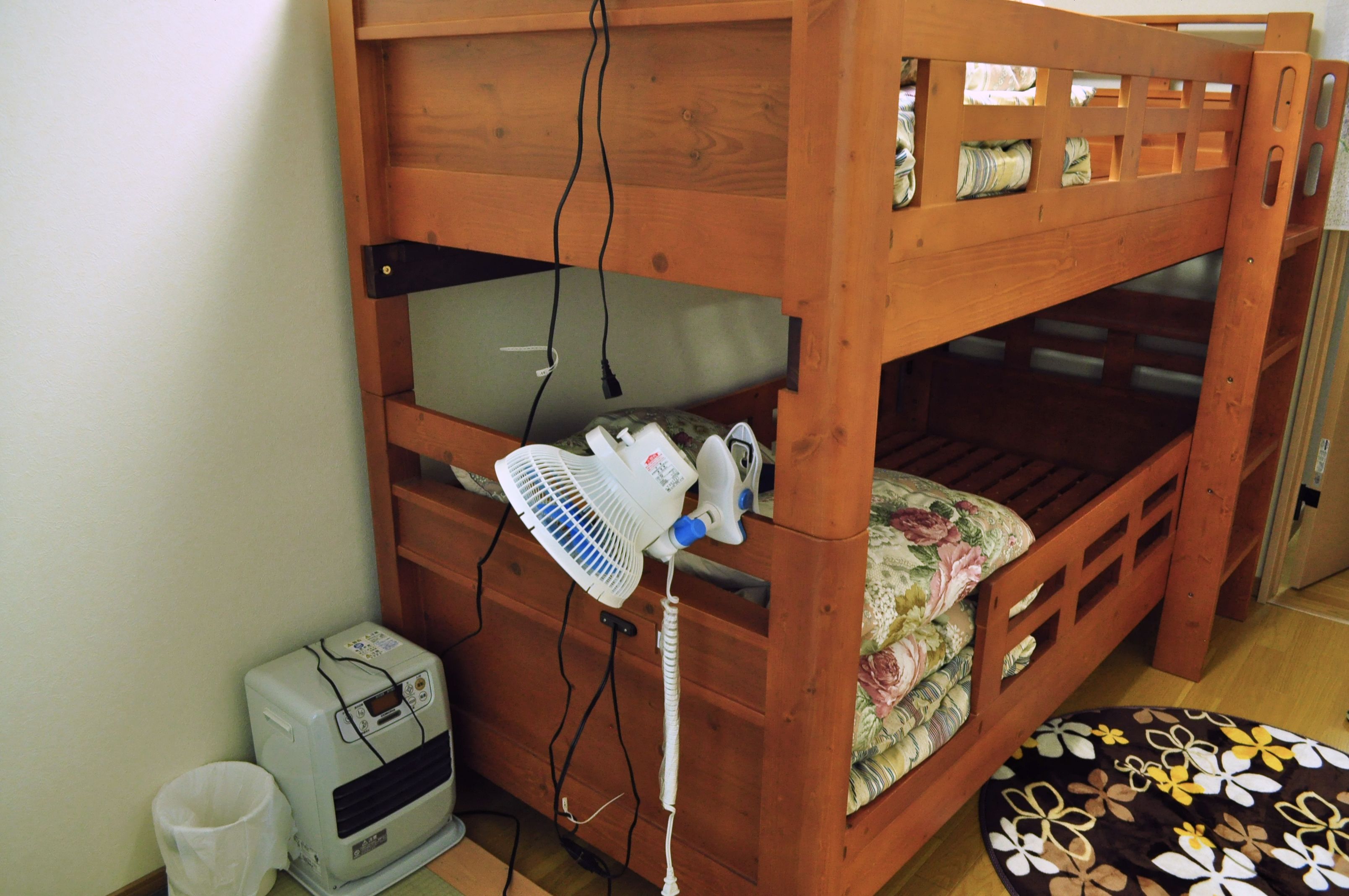 Una cama litera de madera con un ventilador y un calentador en el suelo, un cubo de basura pequeño y una alfombra estampada.