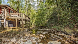 Une cabane dotée d’une terrasse en bois surplombant un ruisseau rocheux, elle-même entourée d’une forêt dense.