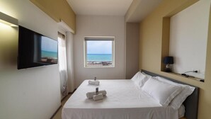Habitación de hotel con cama, televisor montado en la pared, ventana con vista al mar y lámpara en una mesita de noche.
