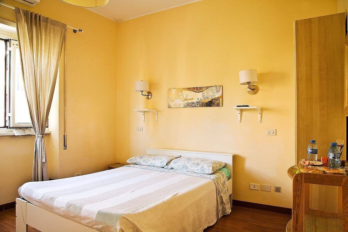 A hotel room with a bed, two wall-mounted lamps, a painting, and a wooden side table with water bottles.