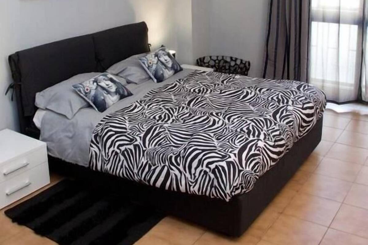 A modern bedroom with a large bed, a nightstand, a decorative wall piece, and a window with curtains.