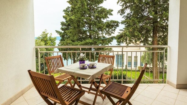 A balcony with a table and four wooden chairs, a potted plant, and a view of trees and a building. - Apartmani Heta (Crikvenica)