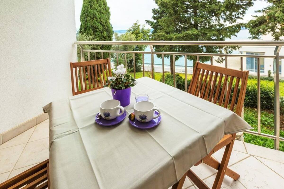 Un balcon avec une table dressée pour deux personnes, un pot de fleurs et une vue sur la verdure et un bâtiment.