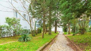A stone pathway leading through a garden with trees and a white building on one side. - Apartmani Heta (Crikvenica)