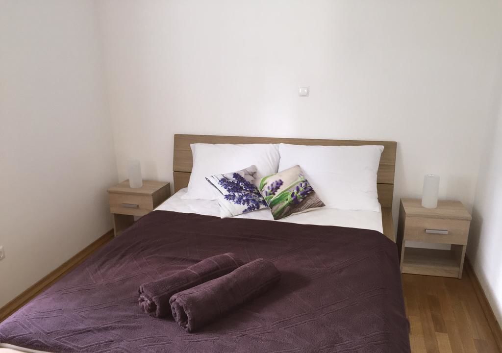 Una cama bien hecha con un edredón púrpura y dos toallas enrolladas, un cabecero de madera y dos mesitas de noche con lámparas.