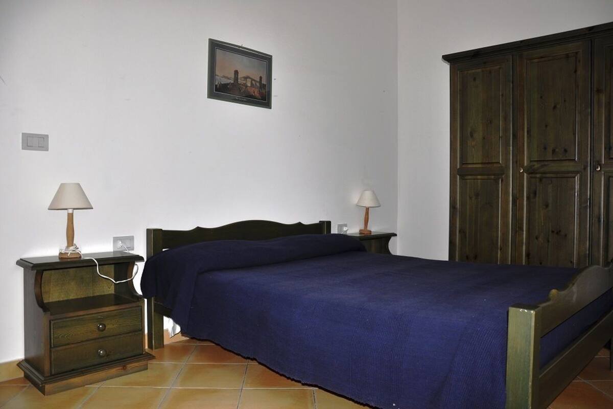 A bedroom with a bed, a wooden wardrobe, a nightstand with a lamp, and a wall-mounted picture.