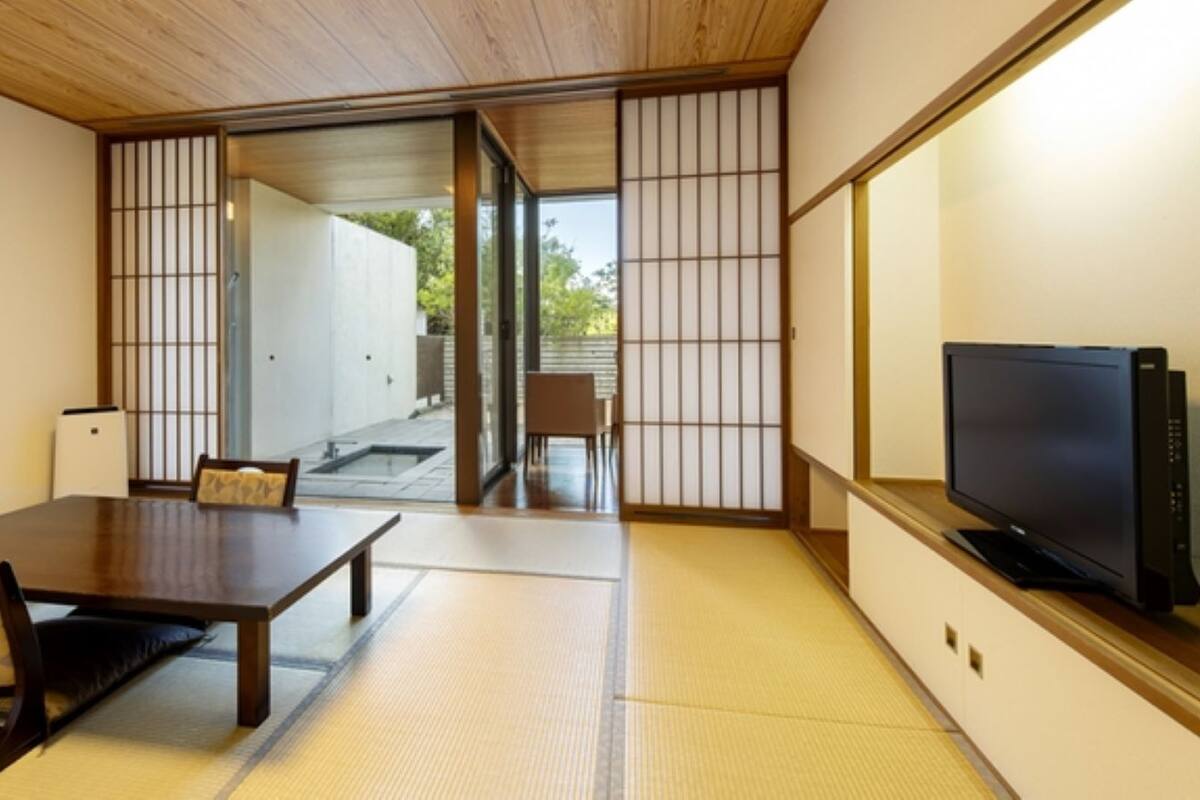 Ein traditioneller japanischer Raum mit Tatami-Böden, einem niedrigen Holztisch, einem Fernseher und Schiebetüren zu einem Außenbereich.
