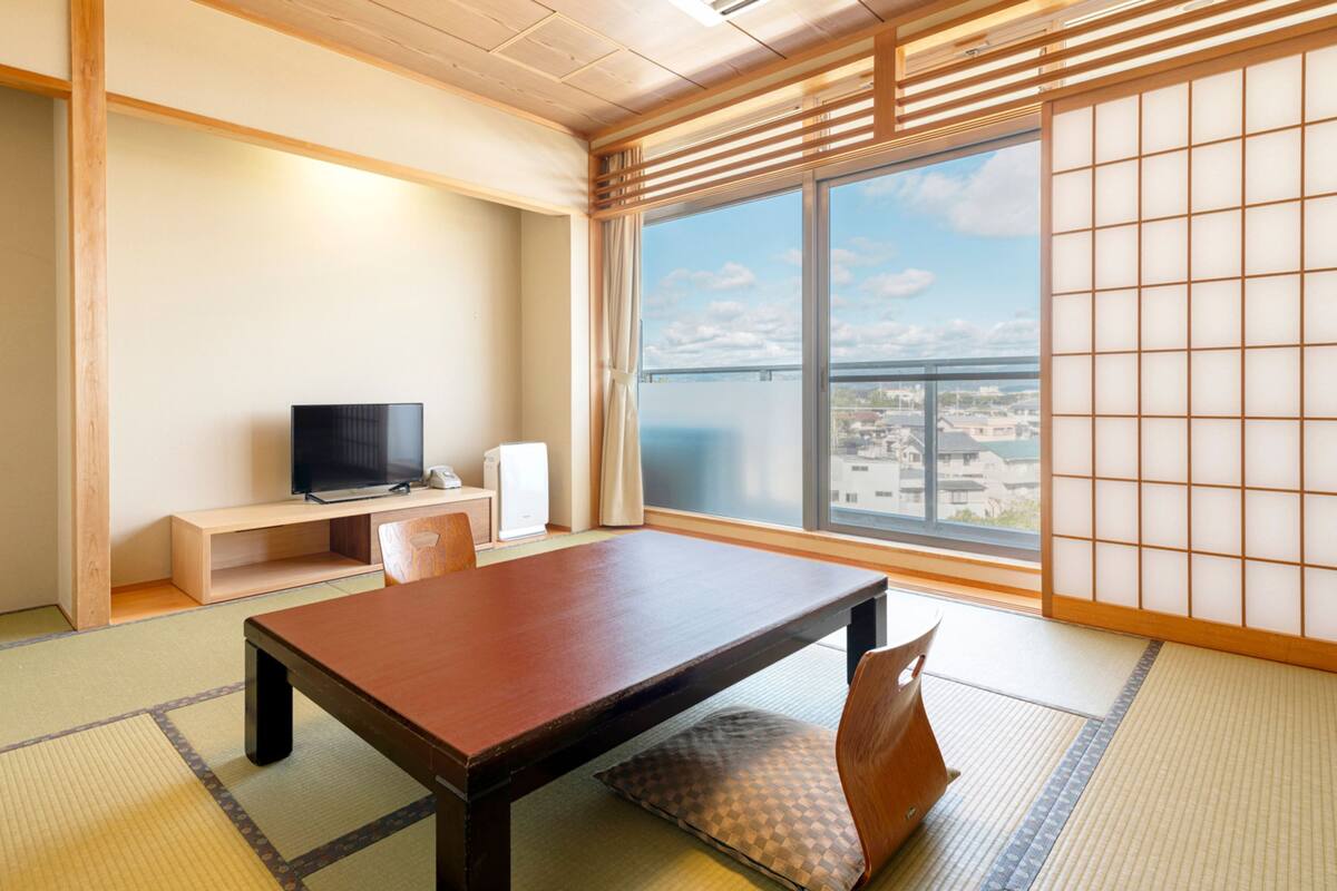 Ein traditioneller japanischer Raum mit Tatami-Böden, einem niedrigen Tisch, einem Fernseher und Blick auf die Stadt durch große Fenster.