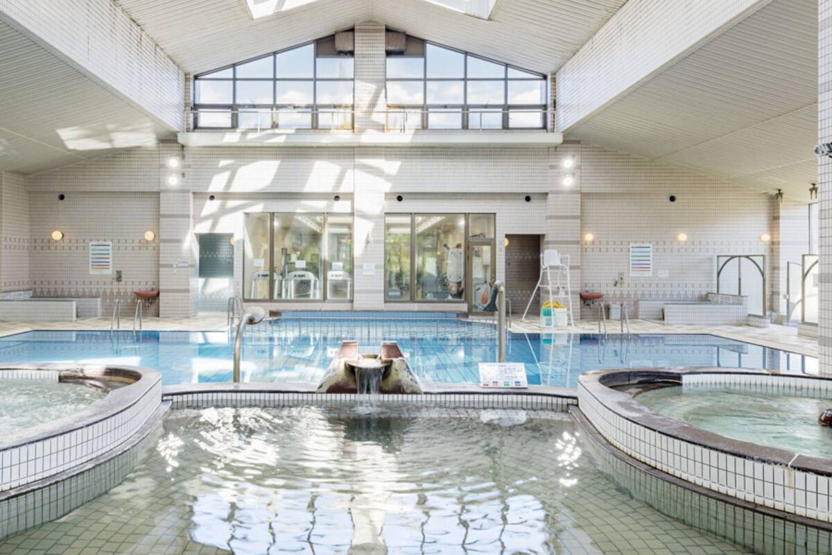 Ein großes Hallenbad mit einem zentralen Whirlpool, umgeben von gefliesten Wänden und einer hohen Decke mit Dachfenstern.