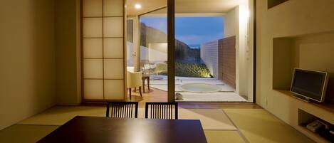 Um quarto tradicional em estilo japonês, com tatames, uma porta deslizante para área externa, uma televisão em uma estante e uma pequena mesa com cadeiras.