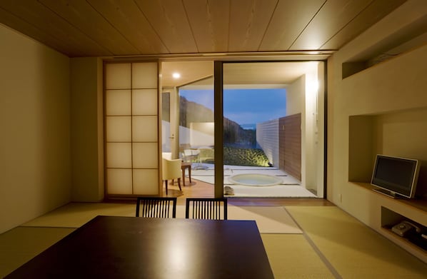 A traditional Japanese-style room with tatami flooring, a sliding door leading to an outdoor area, a television on a shelf, and a small table with chairs.