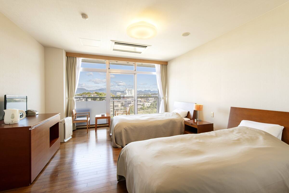 Ein Hotelzimmer mit zwei Betten, einem Schreibtisch mit Lampe, einem Sessel und einem großen Fenster mit Blick auf die Stadt.