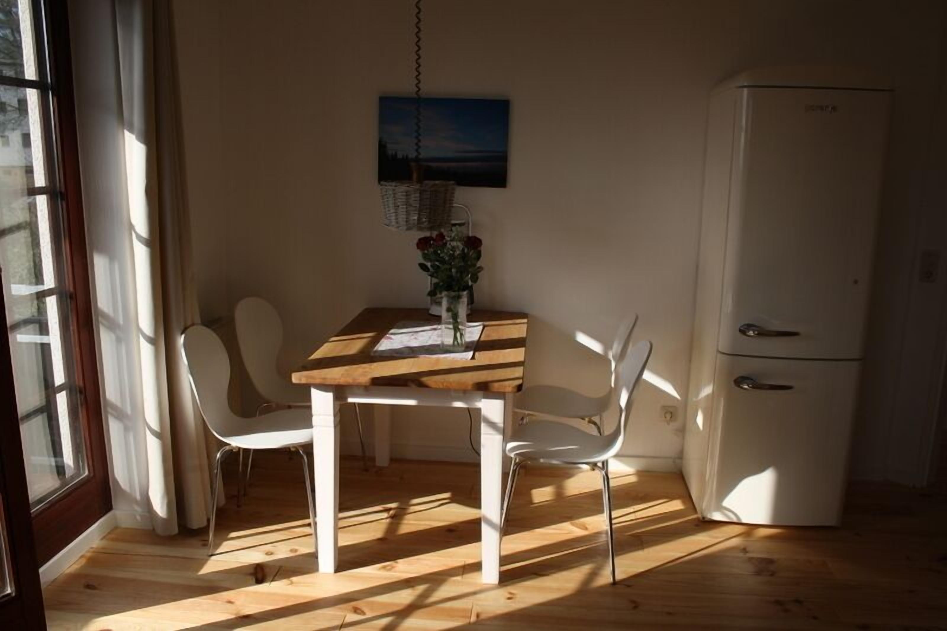 A dining area with a wooden table and white chairs, a white refrigerator, and a window with curtains.