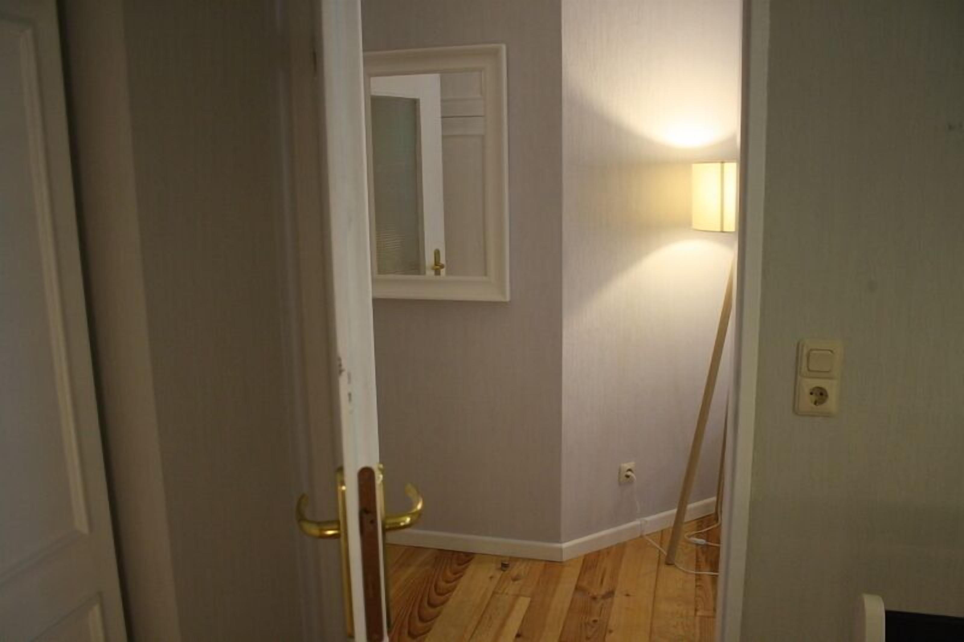 A room with a wooden floor, a white door, a mirror on the wall, and a standing lamp.