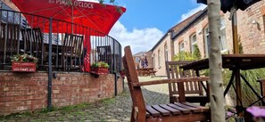A red umbrella with 'PIMMS' branding, a brick wall, and a wooden bench. - The Cross Keys (Knutsford)