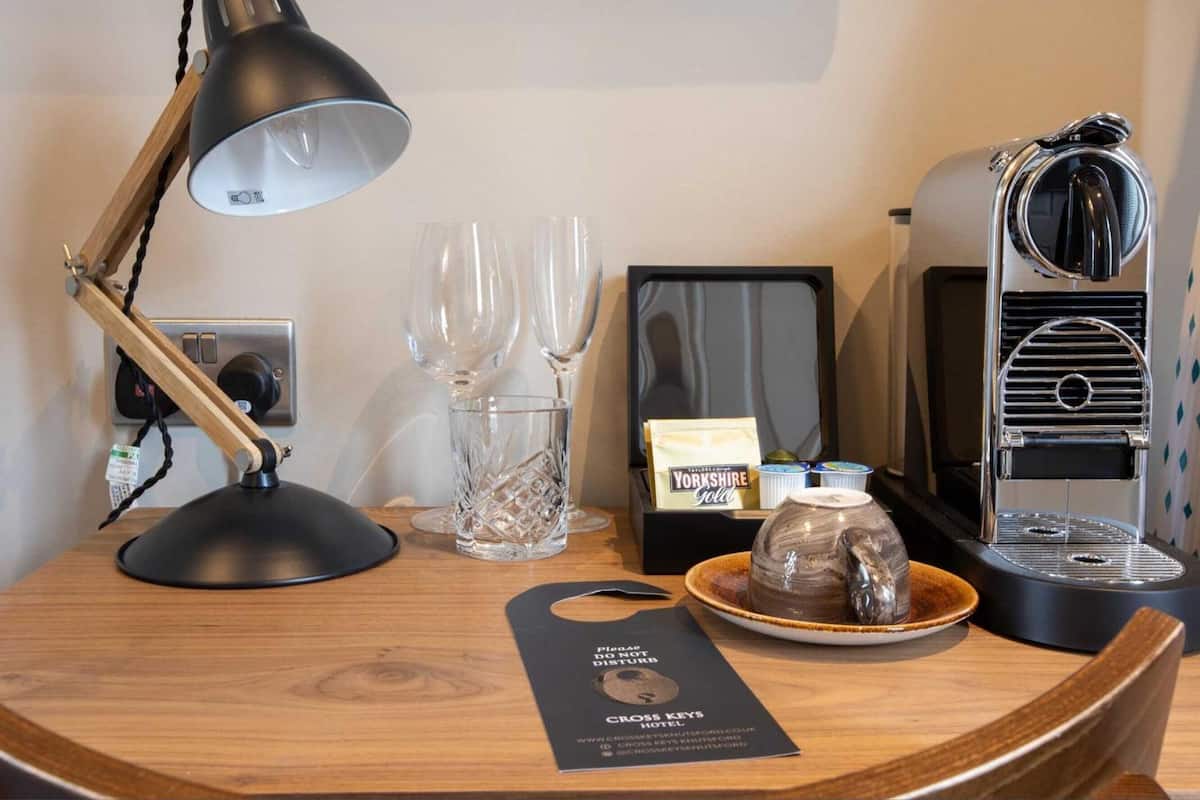 Hotellihuoneen työpöytä, jossa on työpöytälamppu, kaksi viinilasia, kahvinkeitin ja teekuppisetti.