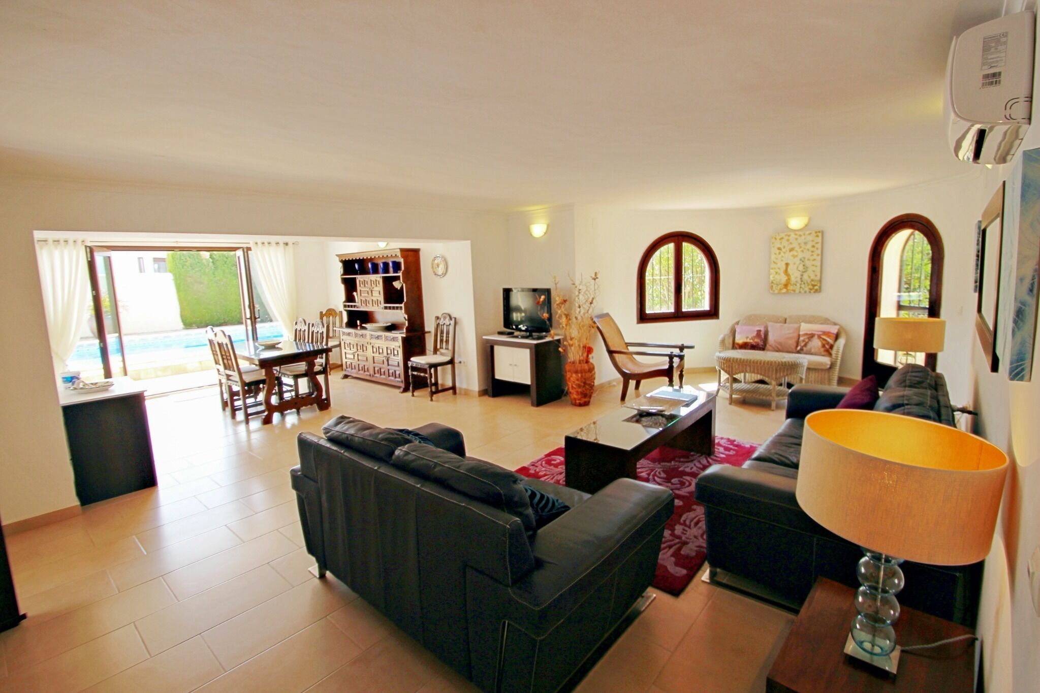 A spacious living room with a sofa, coffee table, and TV. There is a dining area with chairs and a table. The room has large windows and a view of the outdoors.
