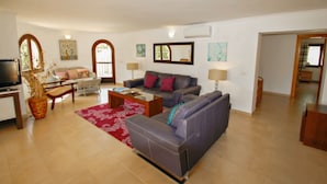 Ein geräumiges Wohnzimmer mit einer Couch, einem Couchtisch und einem Fernseher. Zwei Fenster mit Vorhängen, ein Kamin und ein Essbereich mit Tisch und Stühlen.