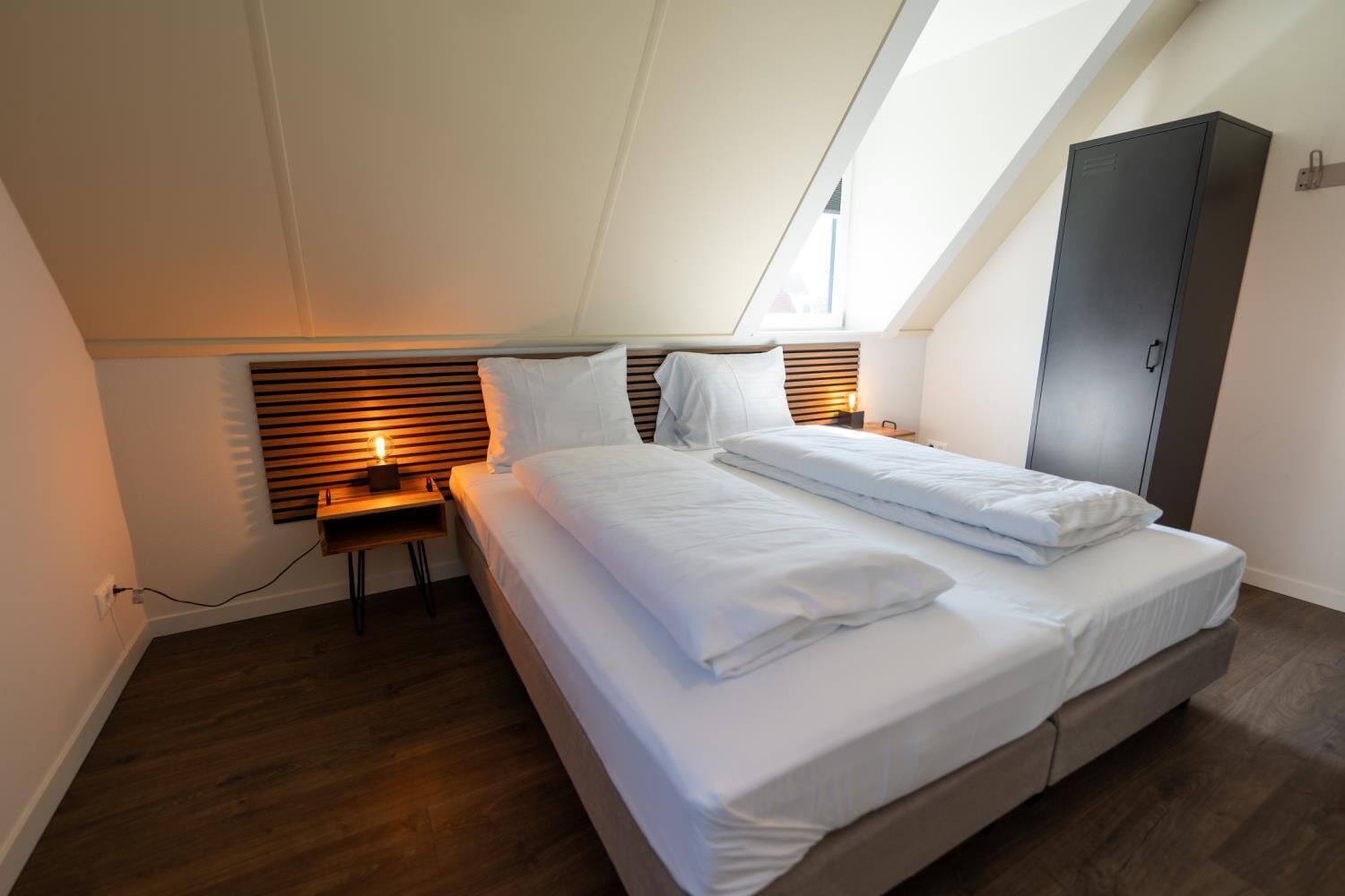 Une chambre à coucher moderne avec un plafond en pente, un lit avec une literie blanche, une table de chevet avec une lampe et une armoire.