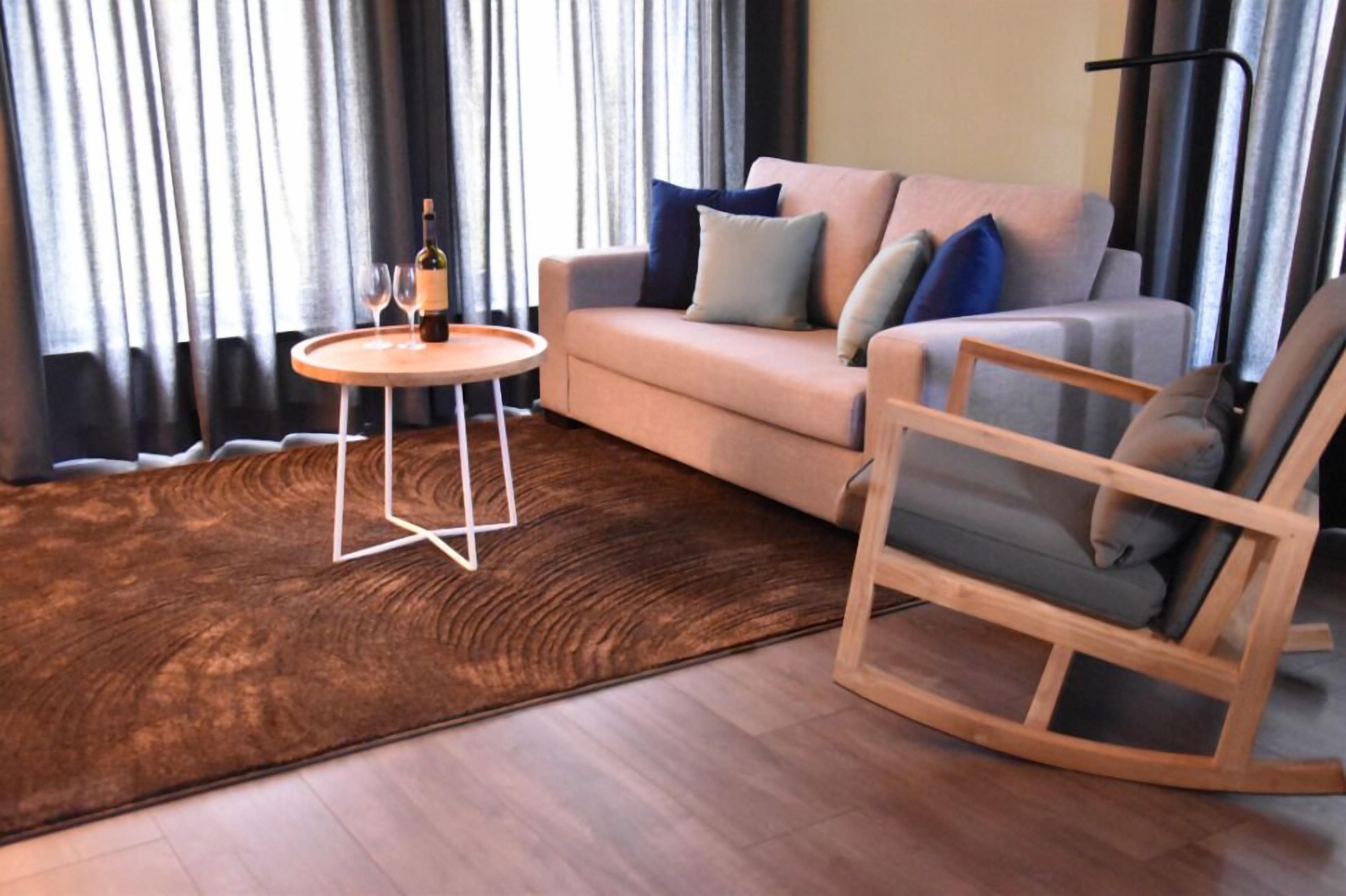 A modern living room with a sofa, a wooden rocking chair, a small round table with a bottle and glasses, and a large brown rug.