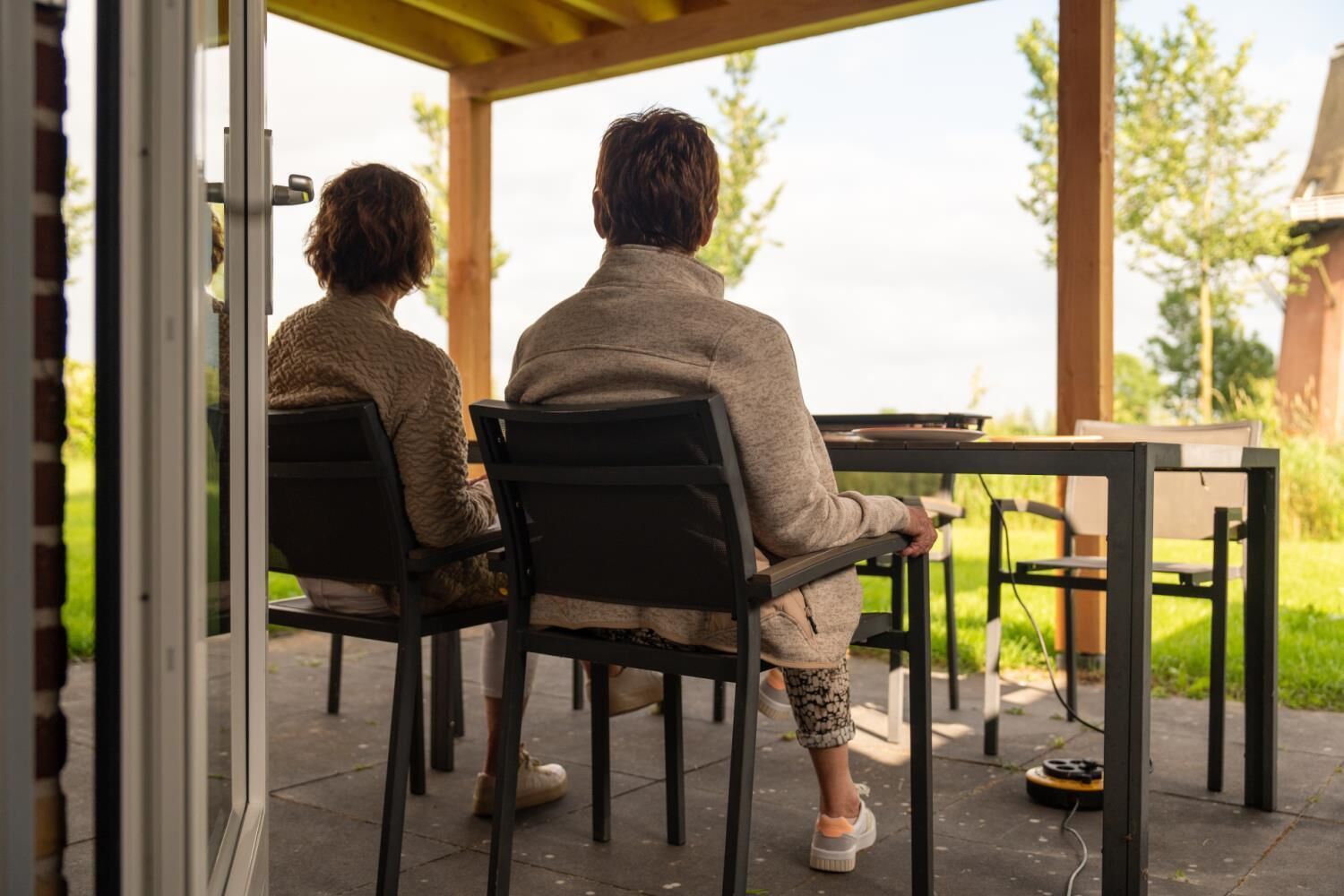 Dos personas sentadas en una mesa en un espacio al aire libre techado, con sillas y una mesa.