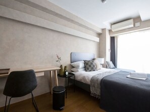 A hotel room with a bed, bedside table, chair, and a small trash bin.