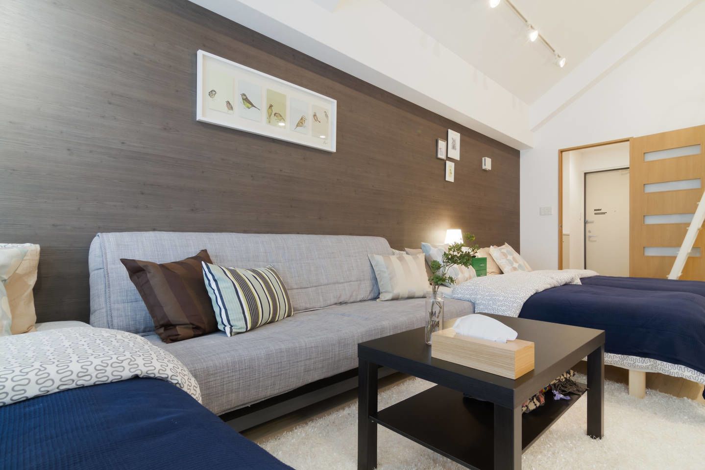 A modern hotel room with a grey sofa, a wooden coffee table, and a bed with a dark blue bedspread.