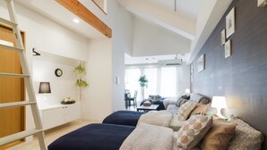 Un dormitorio moderno con dos camas, un escritorio y una silla. Se ve una escalera a la izquierda, una planta en la pared y una ventana al fondo.