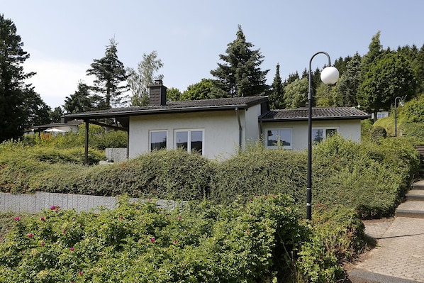 A small, single-story house with a sloped roof, surrounded by dense greenery and a paved walkway. - Landhaus Wald und See (Biersdorf am See)