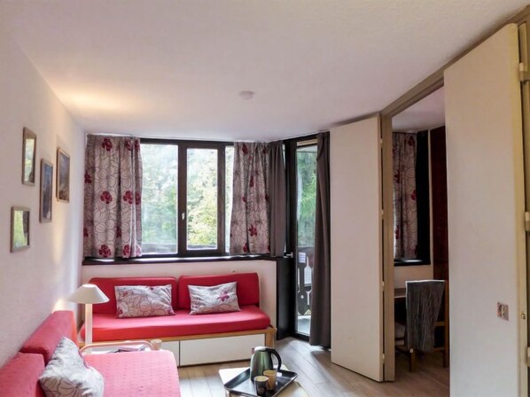 Quarto com sofá vermelho, uma mesinha com chaleira e janela com cortinas florais.