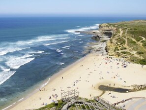 A beach with a rocky cliff, clear blue sea, and a wooden walkway leading to the shore.
