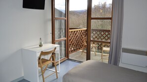 Un dormitorio con cama, escritorio y silla. Se ve una ventana con vista a árboles y montañas.