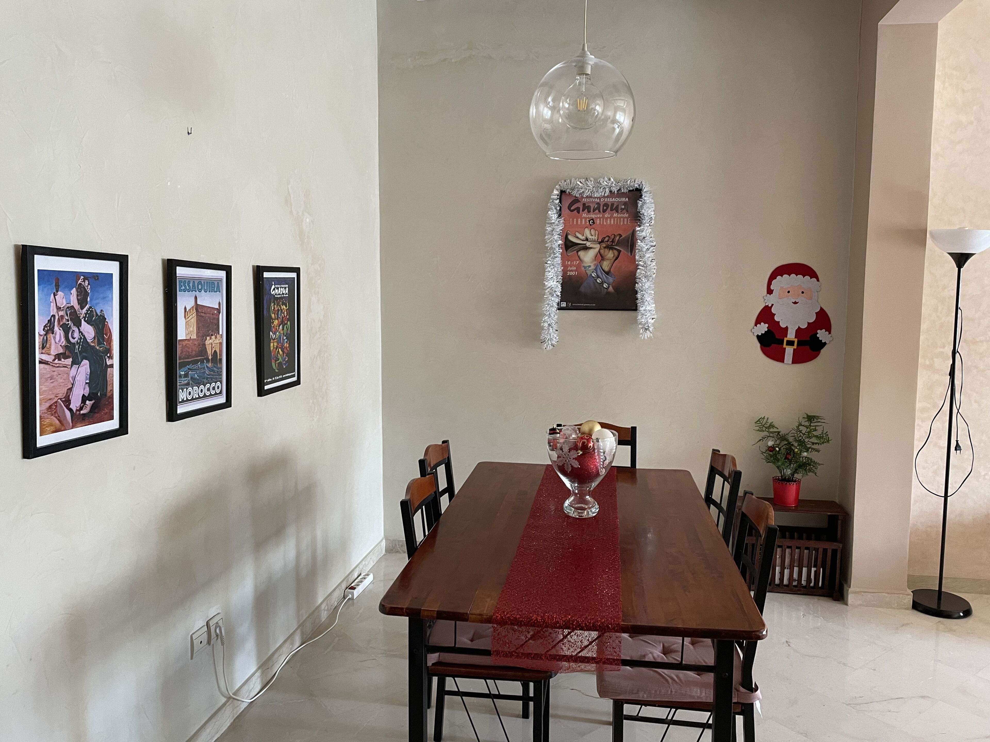 Un coin repas avec une table en bois, des chaises, un chemin de table rouge, un luminaire suspendu, des tableaux encadrés, une affiche et une décoration de Noël.