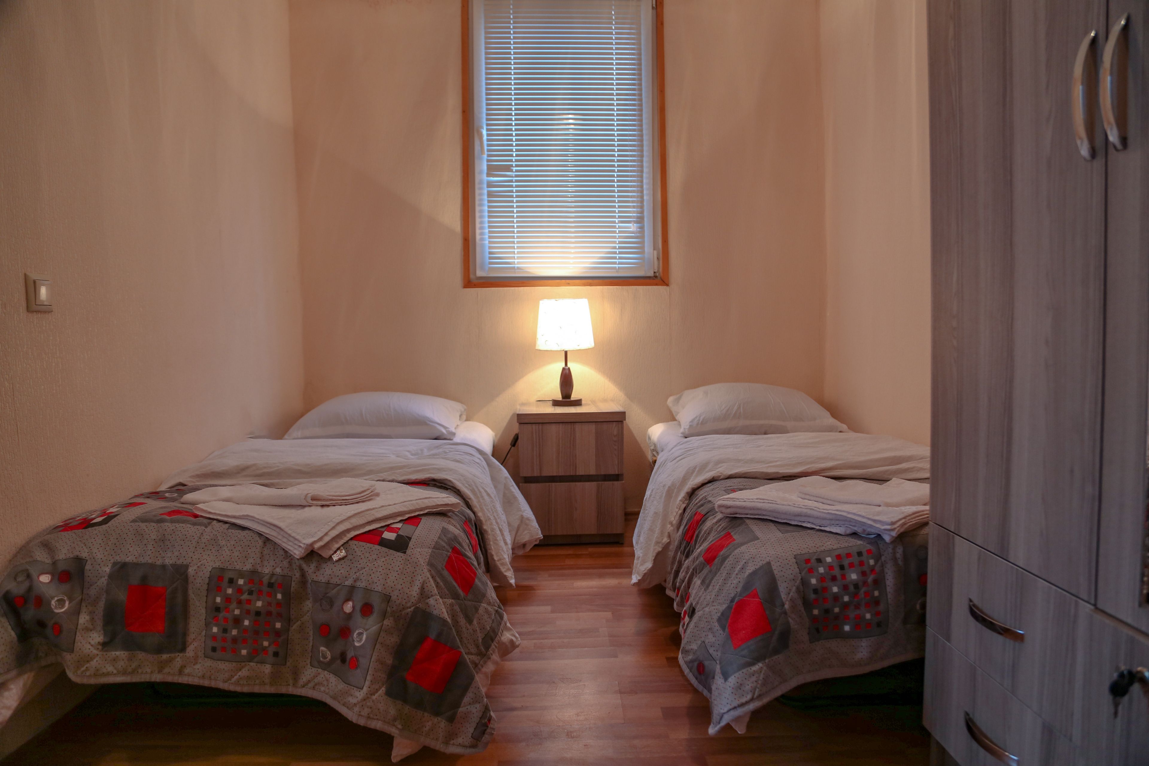 A bedroom with two single beds, a nightstand with a lamp, a window with blinds, and a wardrobe.