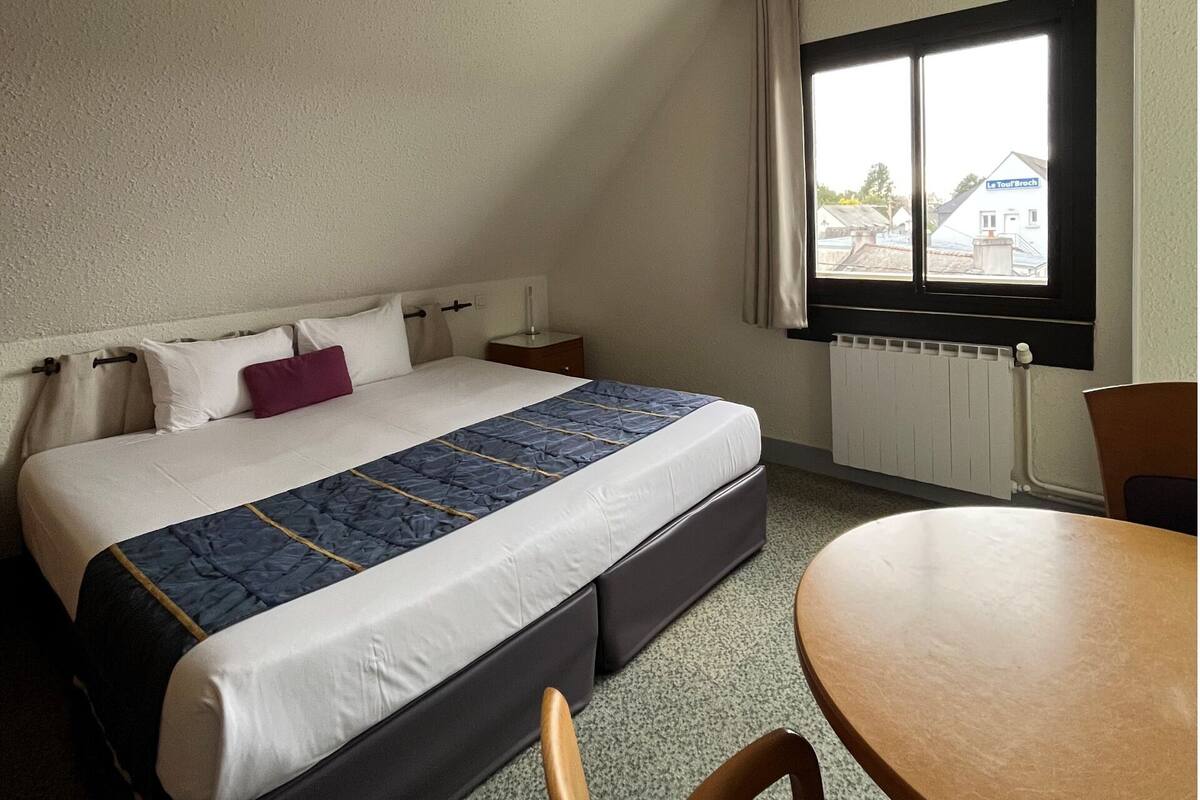 A bedroom with a bed, a window, a radiator, a table, and a chair.