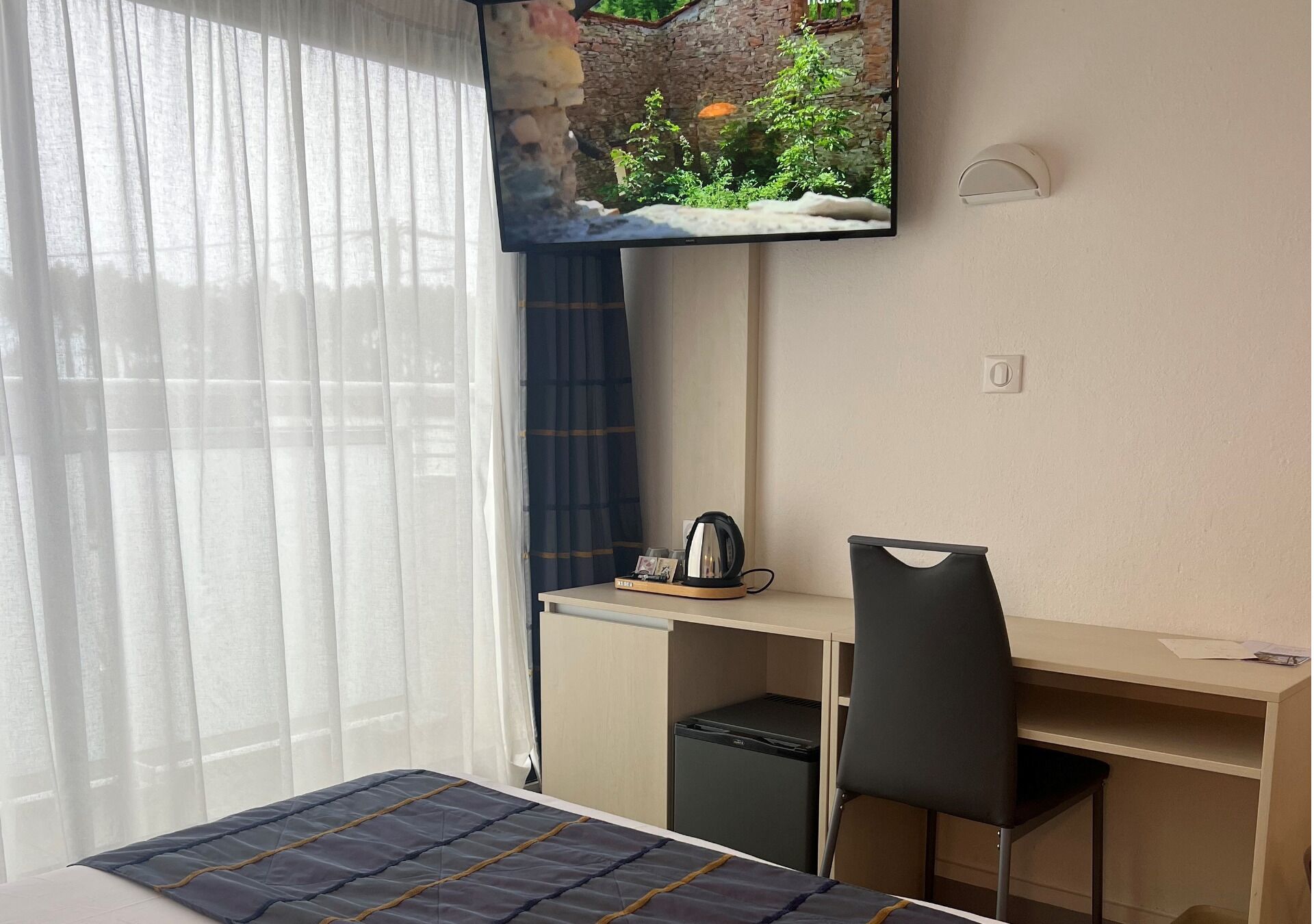 A hotel room with a bed, a desk with a chair, a television mounted on the wall, and a small refrigerator.